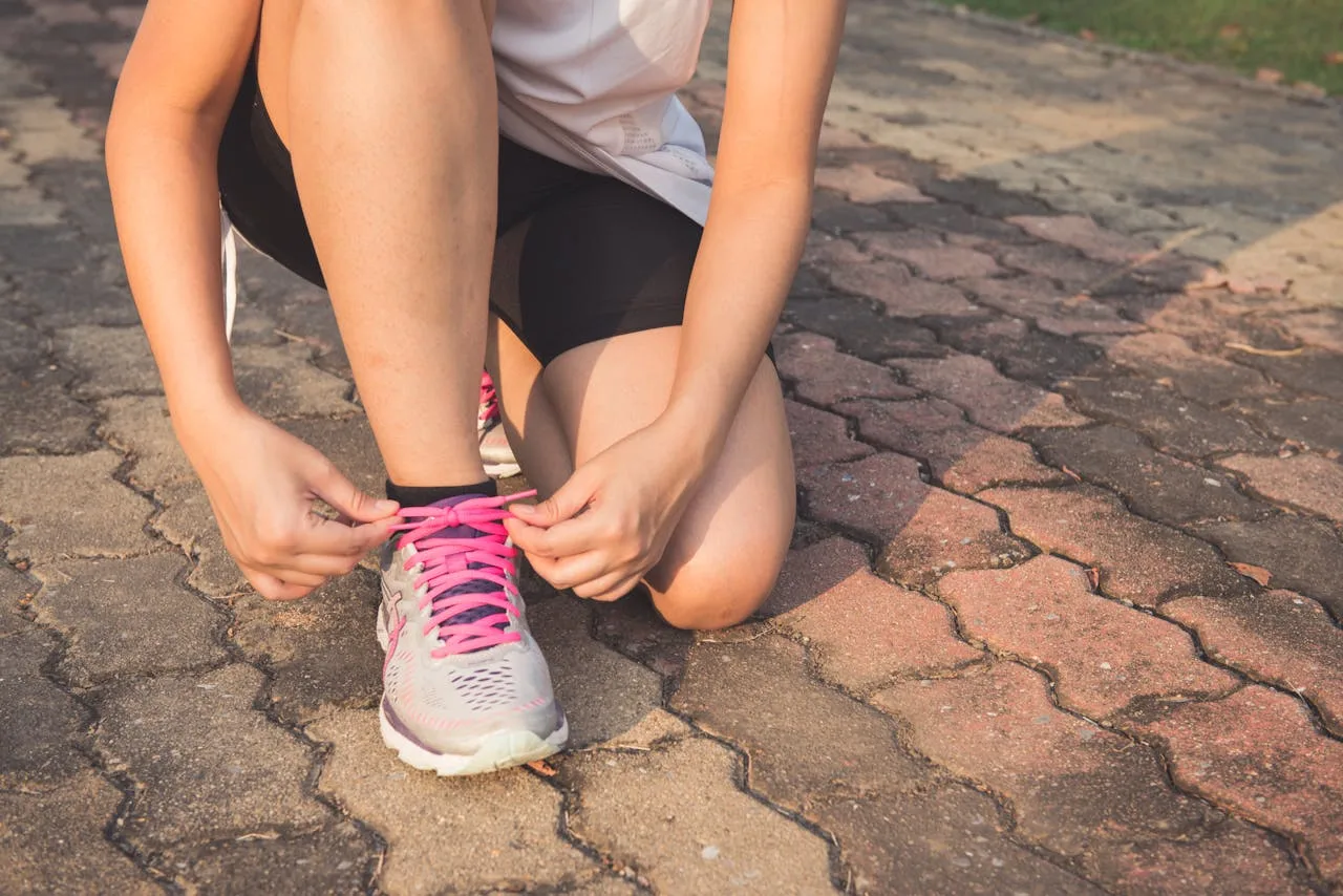 gallery-5 Adult woman tying pink laces on running shoes outdoors. Focuses on fitness and lifestyle.
