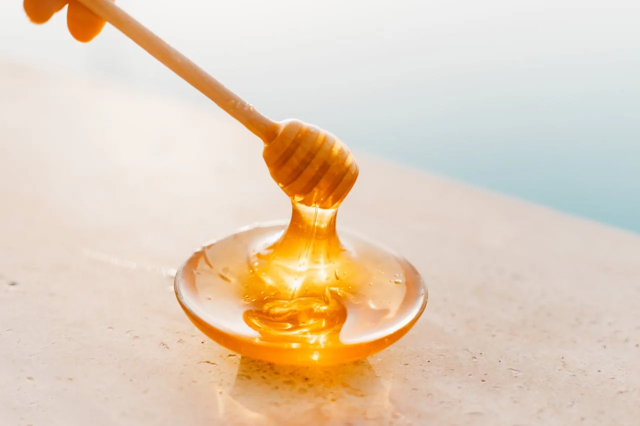 gallery-6 Golden honey flowing from a wooden dipper onto a glass plate, captured in warm daylight.